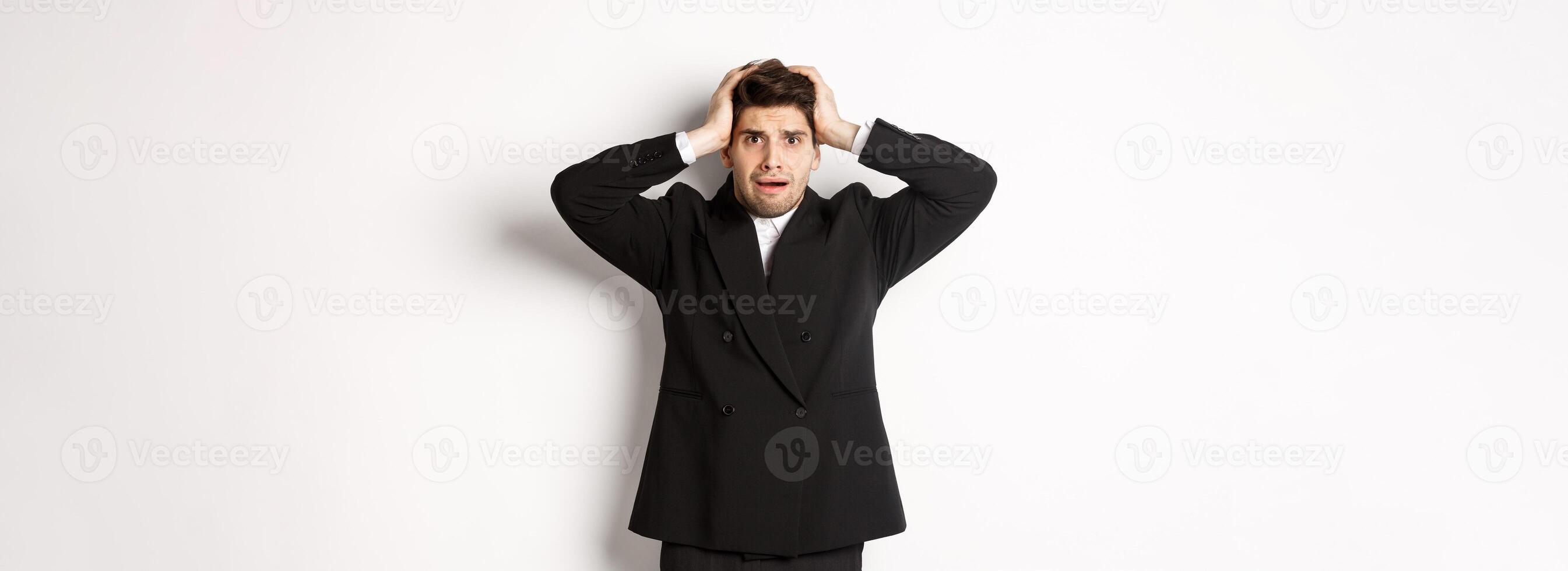 Frustrated and worried businessman in black suit, panicking as looking at trouble, holding hands on head alarmed, standing against white background photo