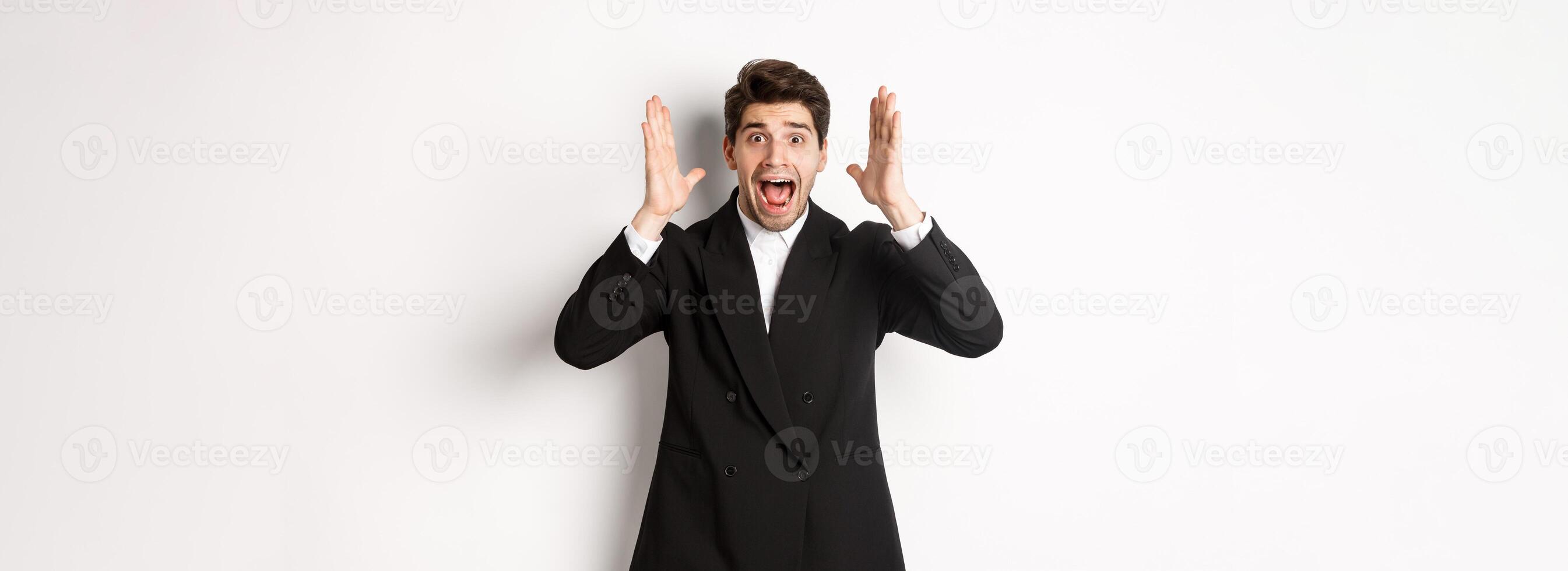 Frustrated and concerned man in black suit, screaming in panic and looking at something shocking, standing over white background photo