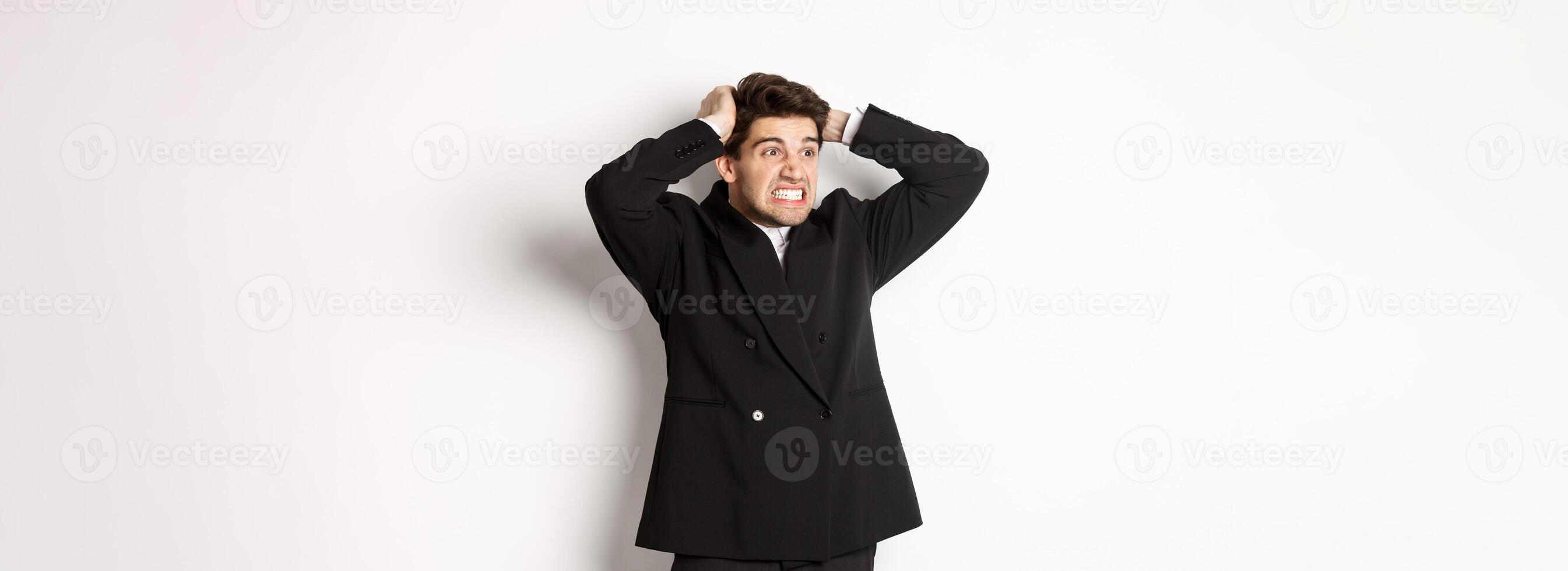 Image of frustrated and angry businessman in black suit, ripping hair on head and grimacing mad, looking left at disaster, standing tensed against white background photo