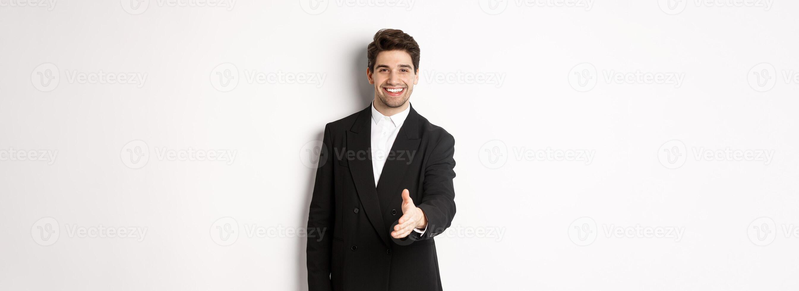 Portrait of handsome businessman in black suit, extending hand for handshake, greet business partners and smiling, welcome to company, standing over white background photo