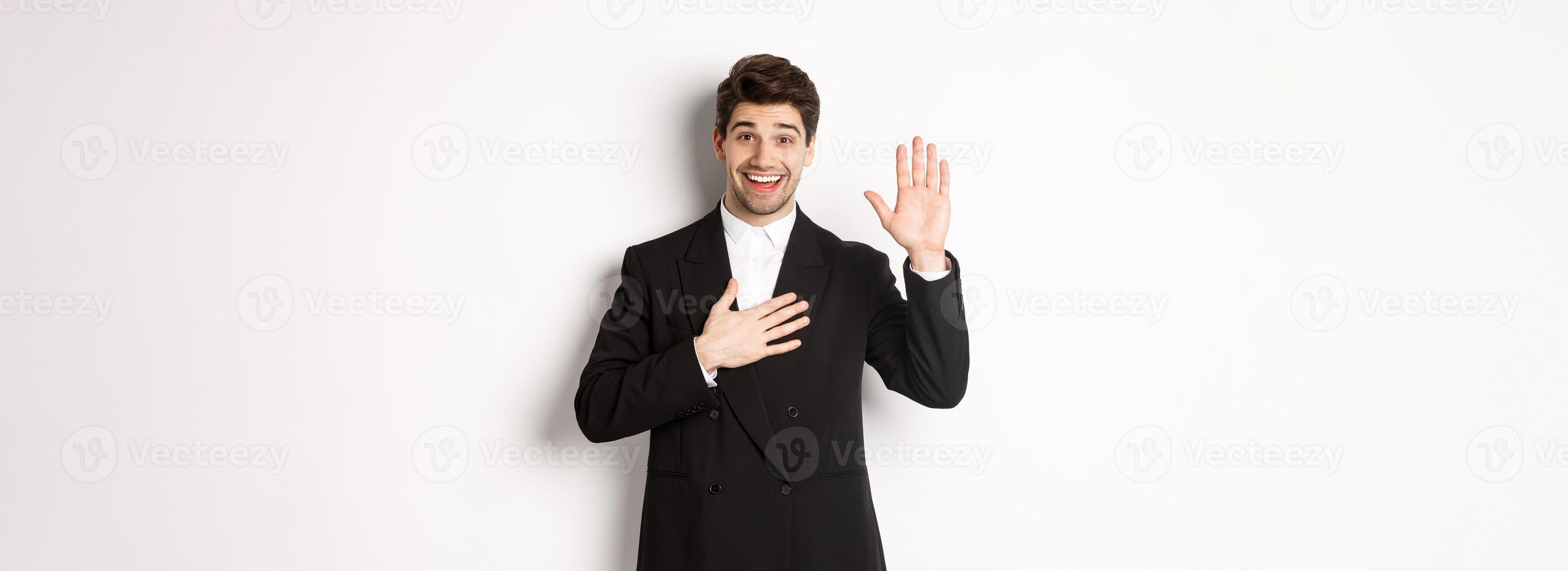 Image of handsome smiling guy telling the truth, raising on arm to promise something, standing over white background in black suit photo