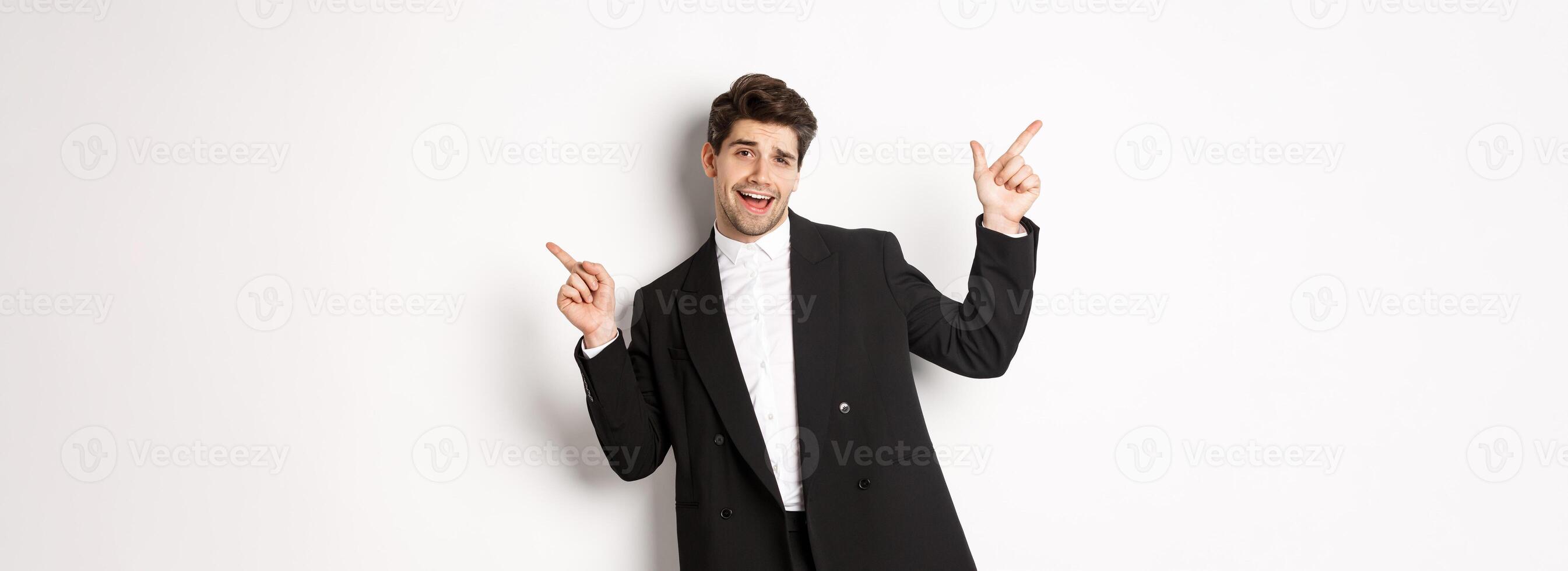 Handsome guy dancing and partying in black suit, pointing fingers sideways, showing two promo banners, standing against white background photo