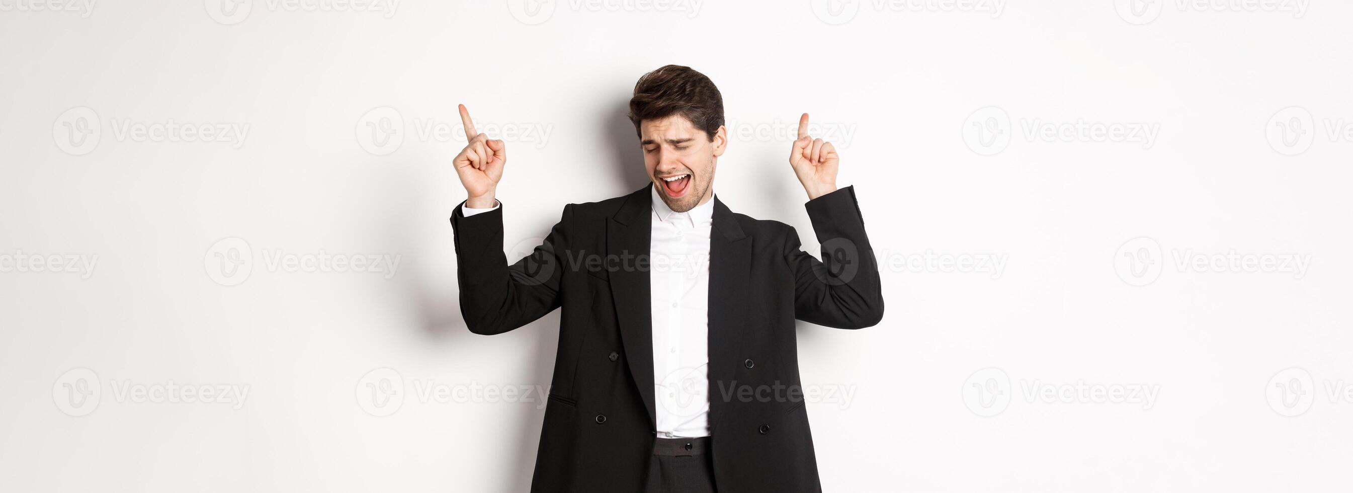 Image of successful handsome businessman dancing, enjoying party, pointing fingers up and having fun, standing over white background, wearing black suit photo