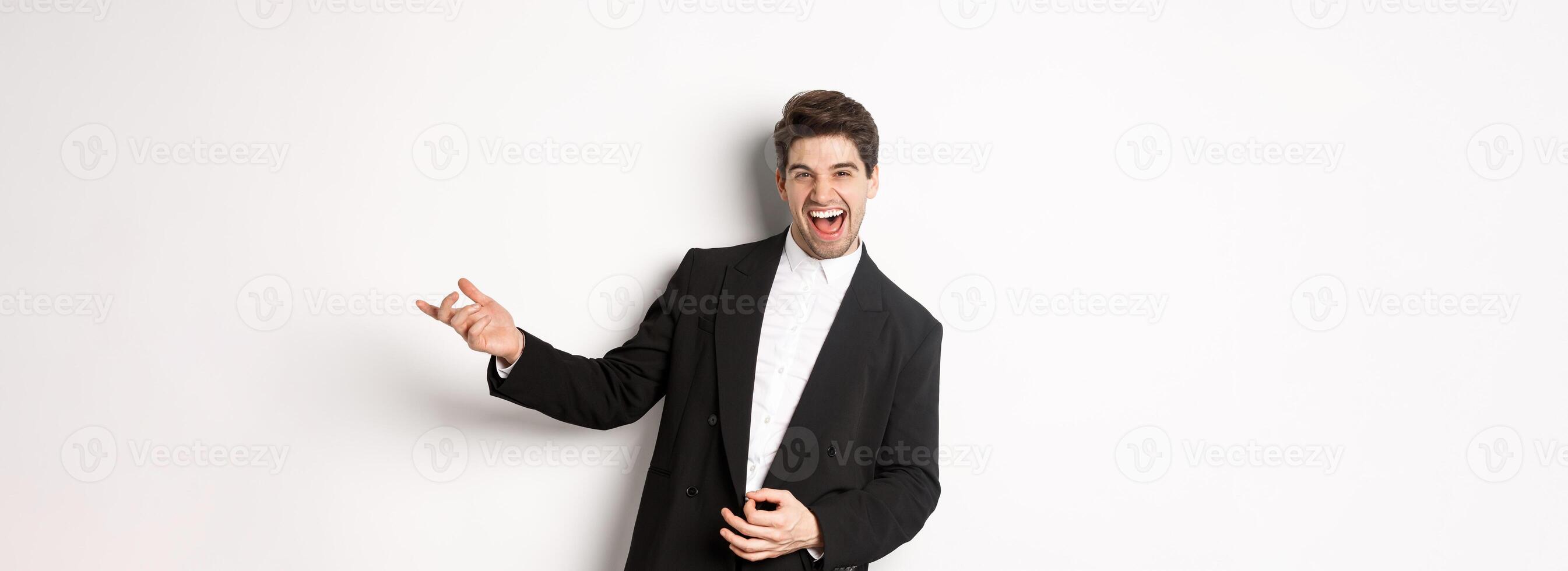 Portrait of happy man dancing at party, playing on invisible guitar and laughing, standing in black suit against white background photo