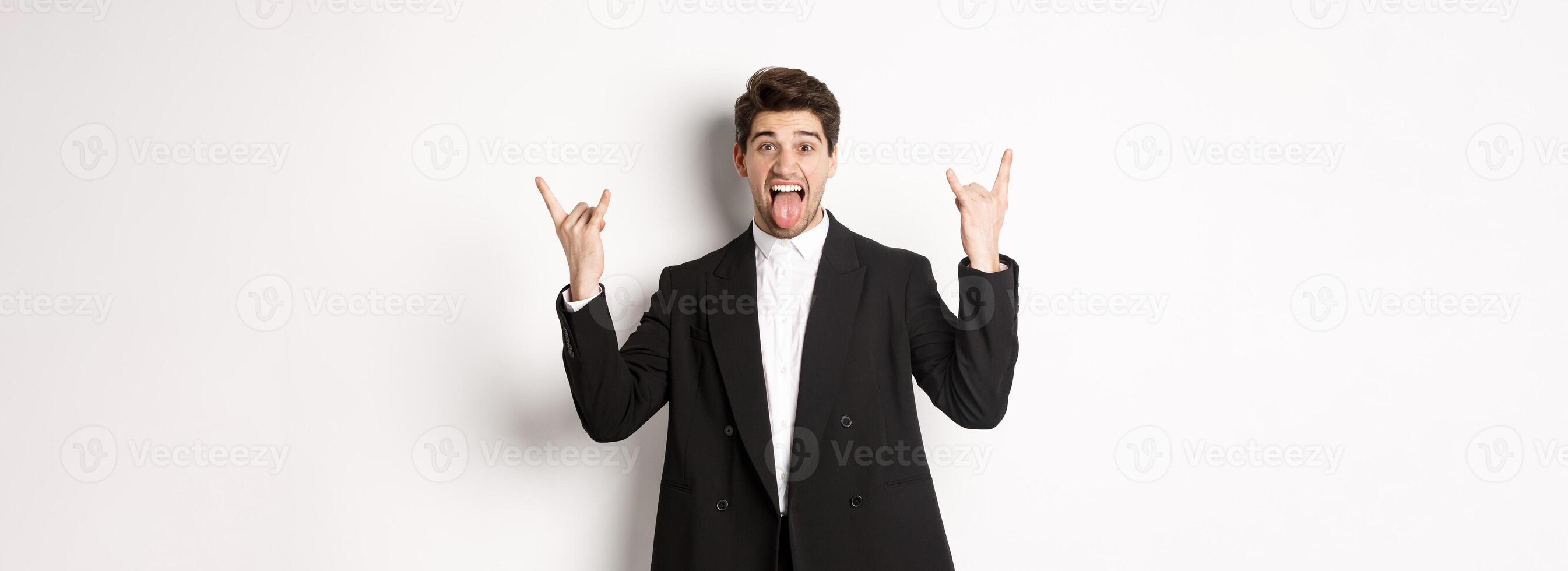Portrait of happy attractive guy having fun at party, wearing black suit, showing rock-n-roll sign and tongue, standing excited against white background photo