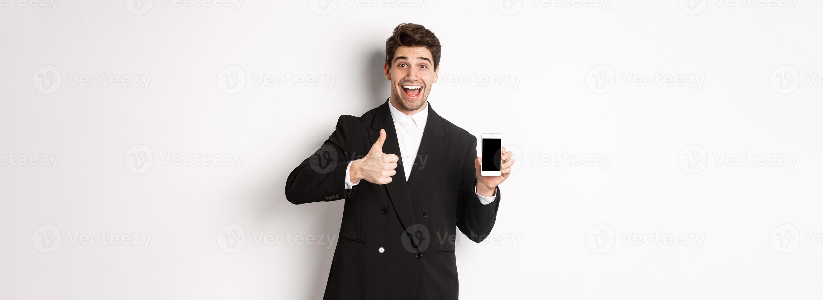 Image of handsome male entrepreneur in black suit, recommending app or online shop, showing thumbs-up and smartphone screen, standing over white background photo