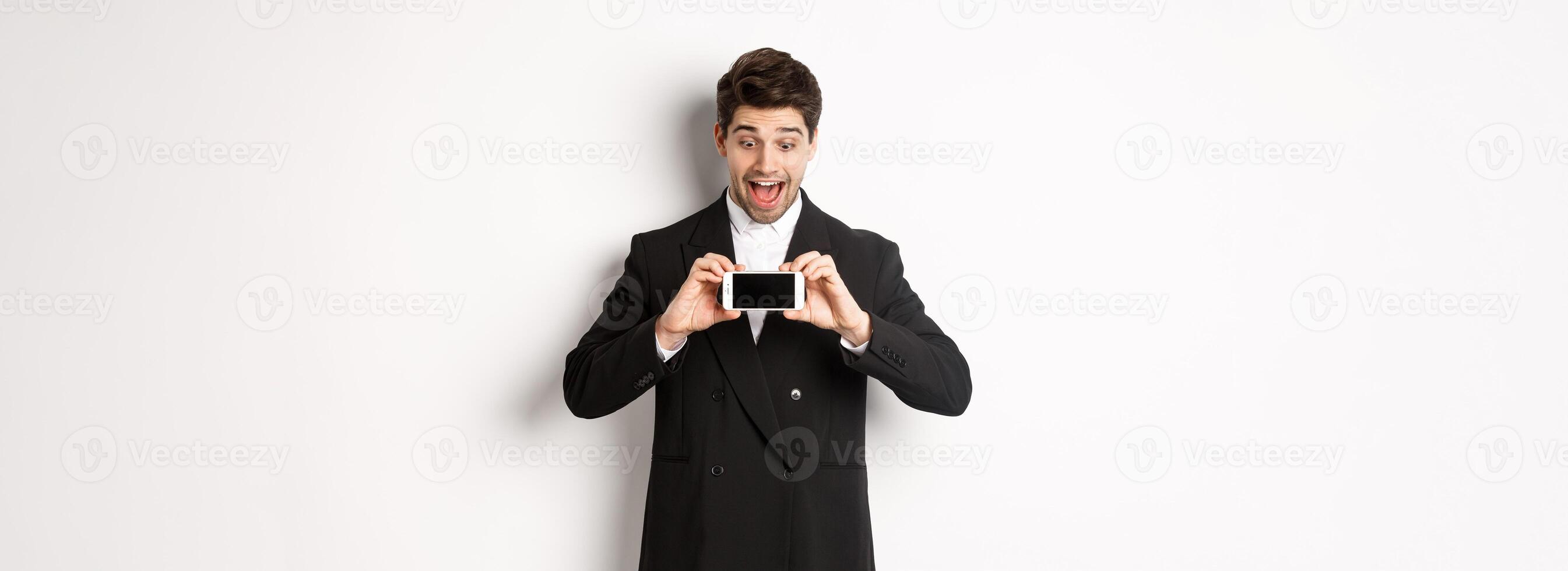 Portrait of excited handsome man showing smartphone screen, looking amazed at display with advertisement, standing over white background in black suit photo