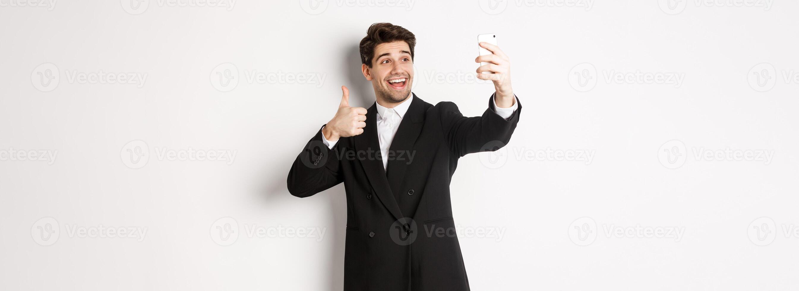 Portrait of good-looking man taking selfie on new year party, wearing suit, taking photo on smartphone and showing thumbs-up, standing against white background