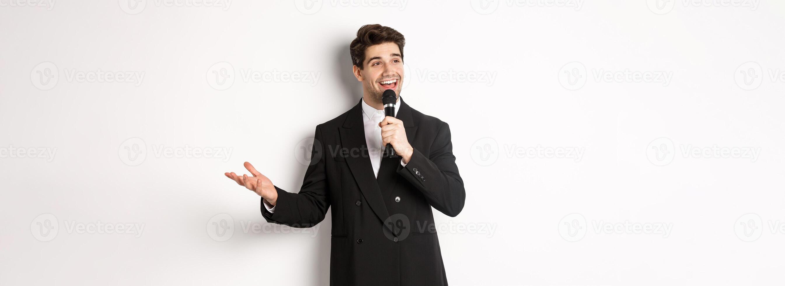 Portrait of handsome man in black suit singing a song, holding microphone and giving speech, standing against white background photo