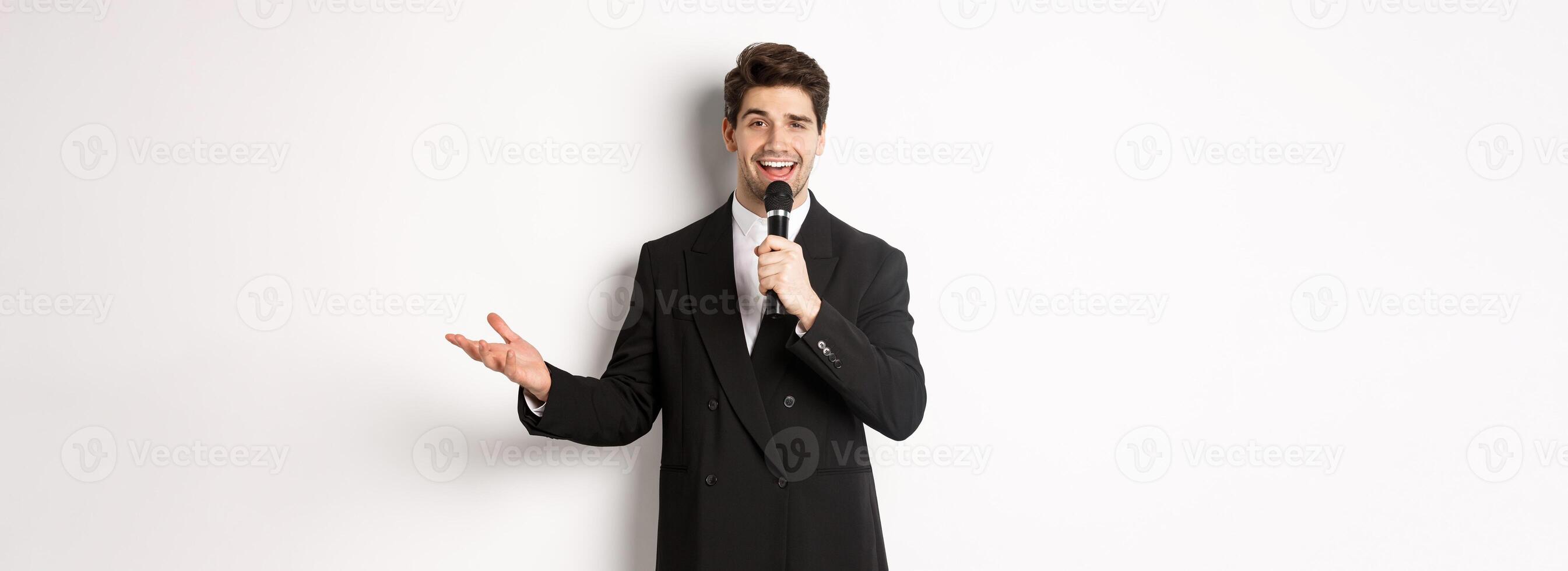 Portrait of handsome man in black suit singing a song, holding microphone and giving speech, standing against white background photo