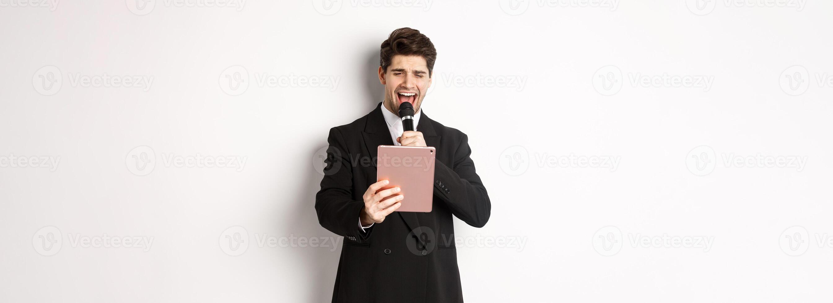 Image of handsome man in black suit, singing karaoke on digital tablet, holding microphone, standing over white background photo