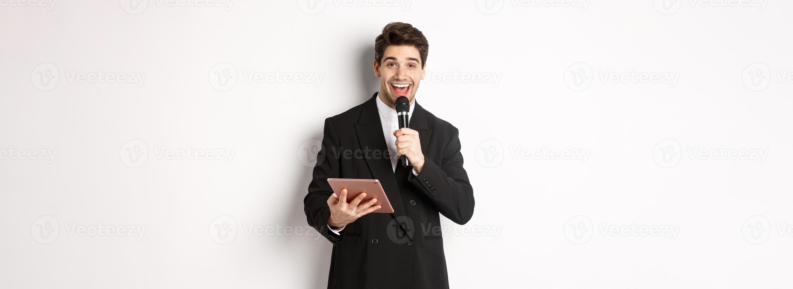 Image of handsome male performer, giving a speech, entertain people at party, holding microphone and digital tablet, standing over white background photo