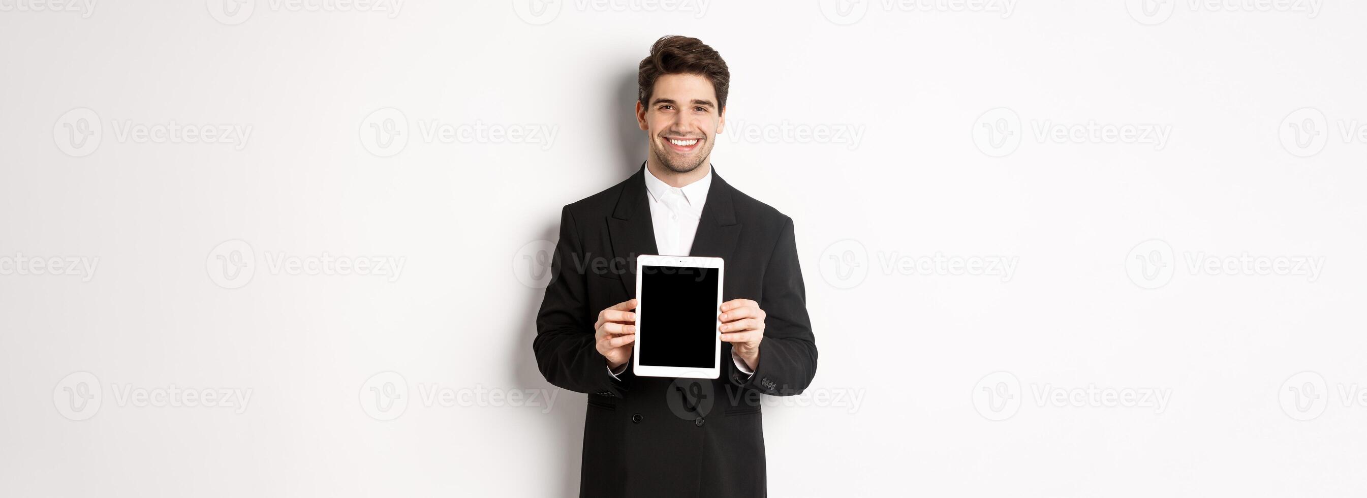 Portrait of handsome businessman in trendy suit, showing digital tablet screen and smiling, standing against white background photo