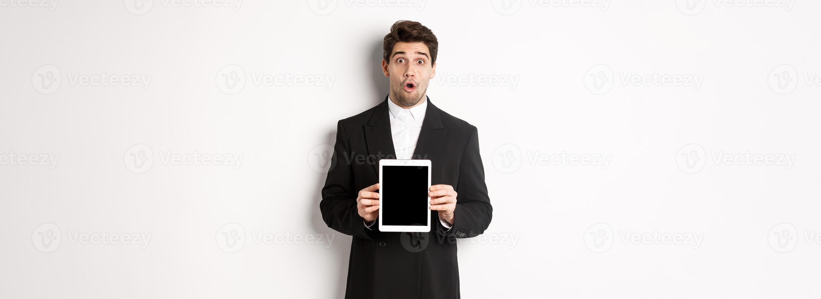 Image of surprised handsome man in black suit, showing digital tablet screen and looking amazed, standing against white background photo