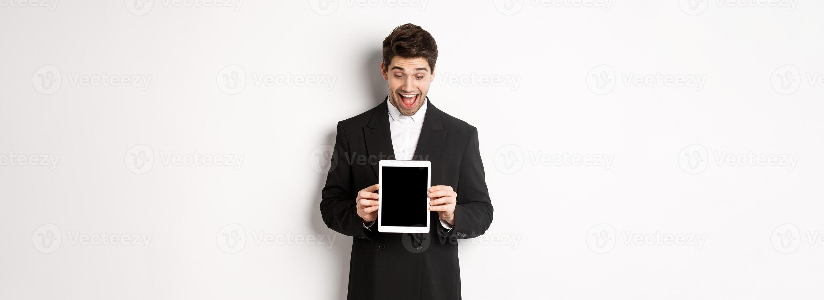 Portrait of amazed handsome man in black suit, showing digital tablet screen with excitement, standing over white background photo