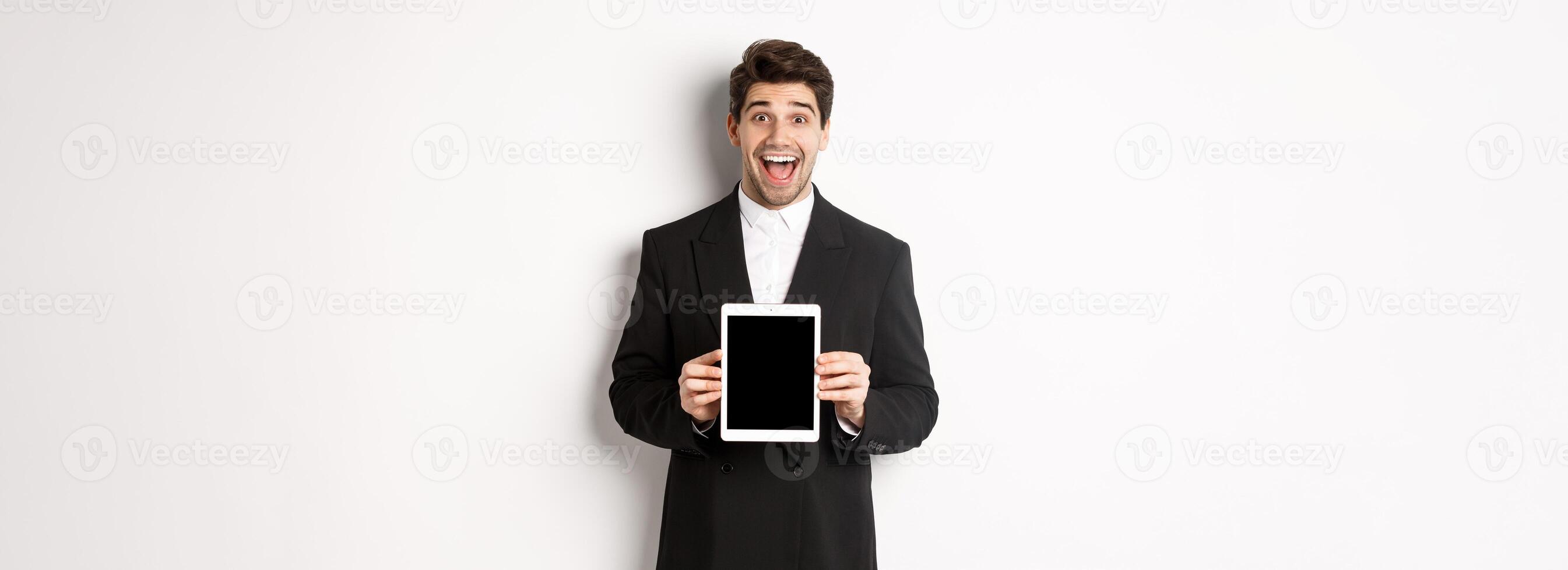 Image of attractive male entrepreneur in trendy suit, showing digital tablet screen and smiling amazed, standing over white background photo