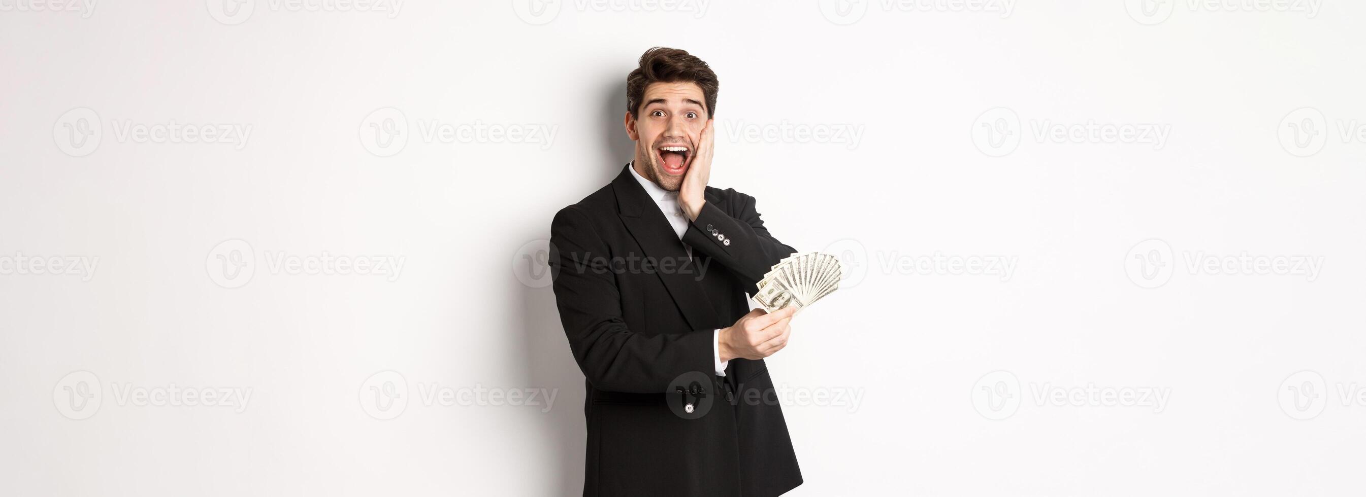 Image of rich and happy man in black suit, winning prize, holding money and looking excited at camera, standing over white background photo