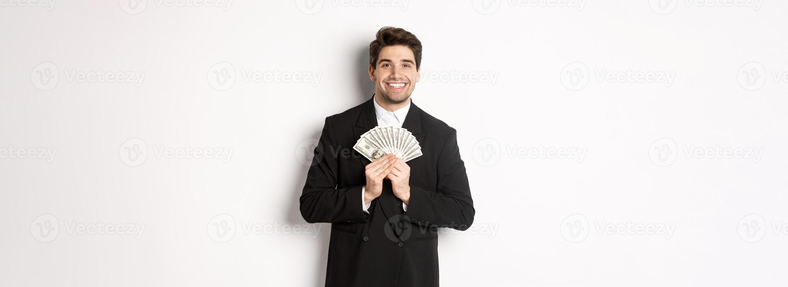 Portrait of happy and pleased handsome man in suit, hugging money and looking satisfied, standing over white background photo