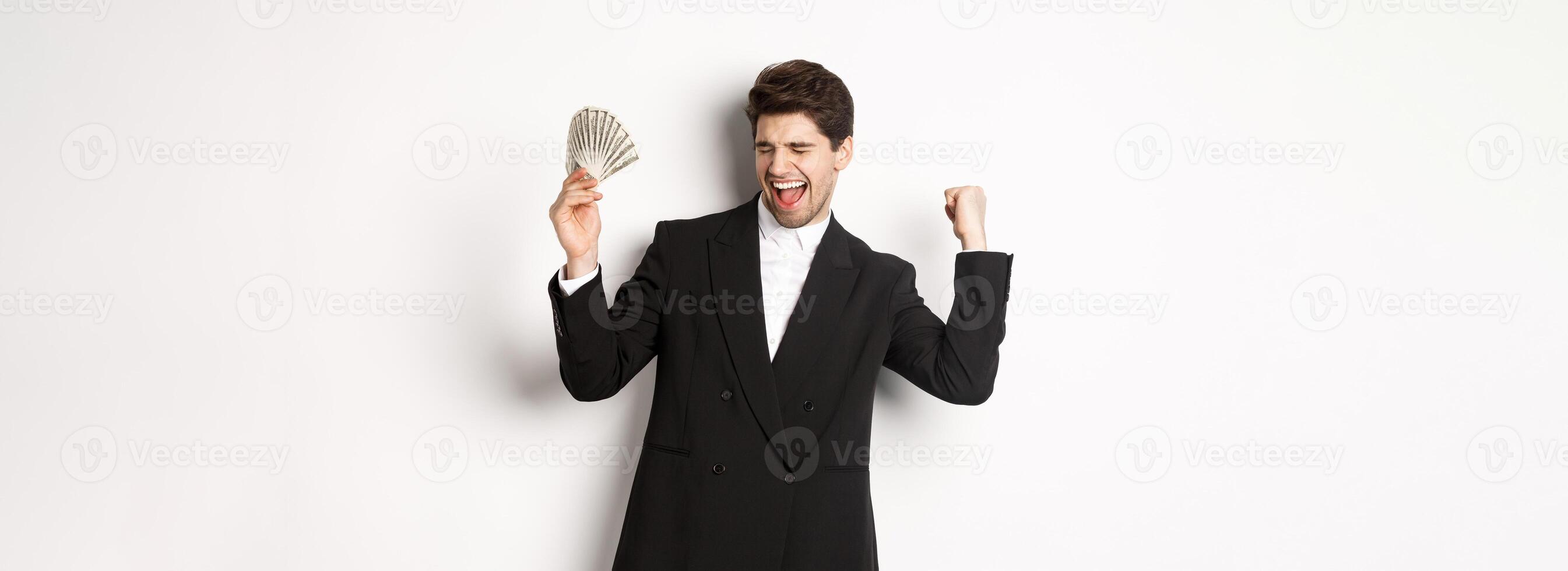 Portrait of handsome and successful businessman in suit, dancing with money, standing against white background photo