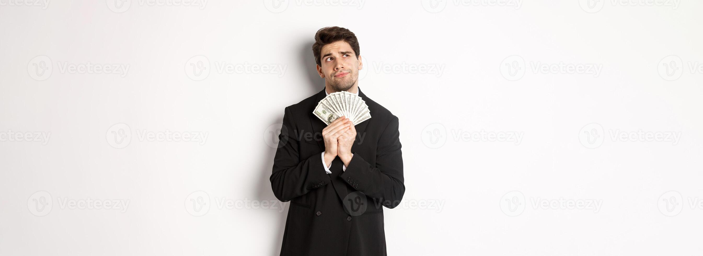 Image of handsome businessman in black suit, holding money and thinking about investment, making shopping plans, standing over white background photo