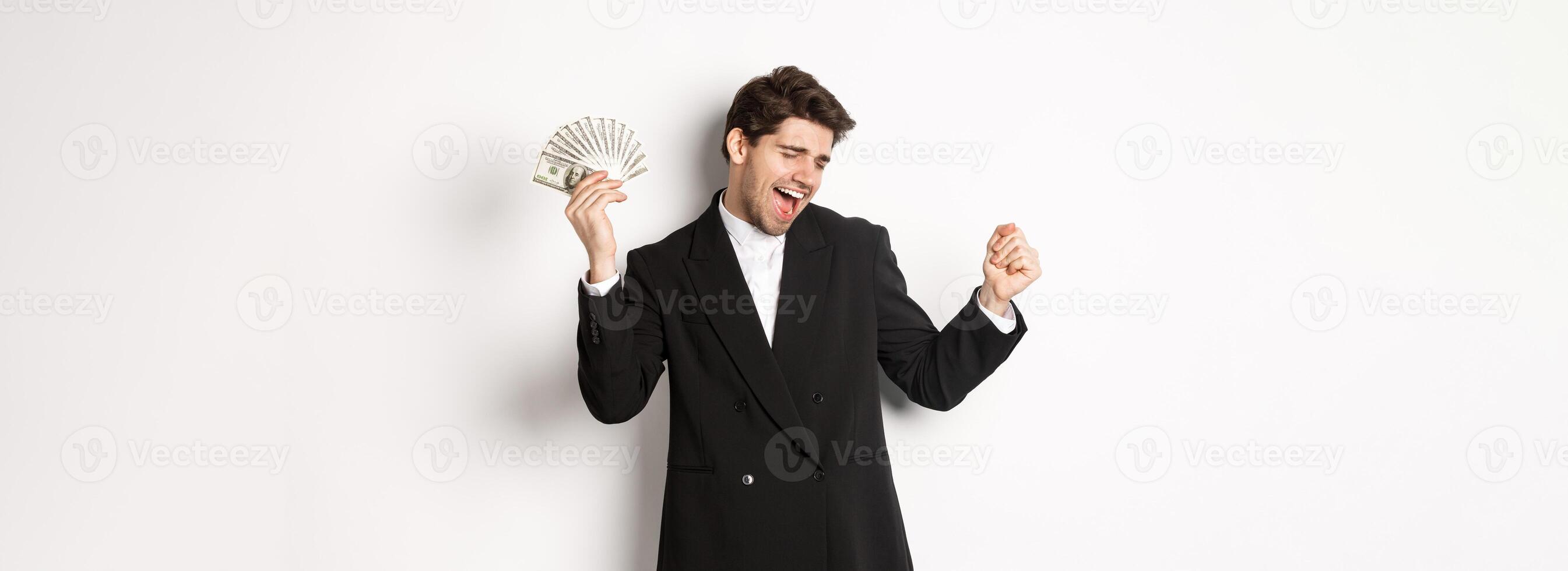 Portrait of handsome and successful businessman in suit, dancing with money, standing against white background photo