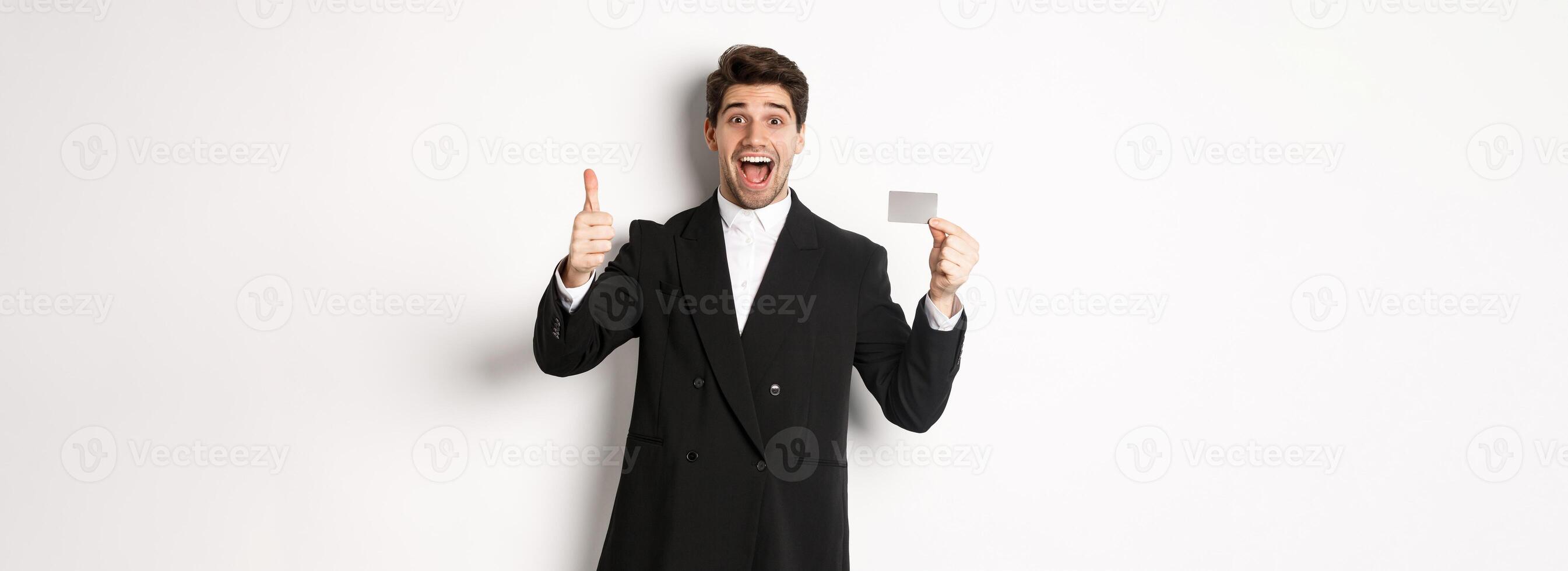 Image of good-looking businessman in black suit, showing thumbs-up and credit card, recommending bank, standing against white background photo