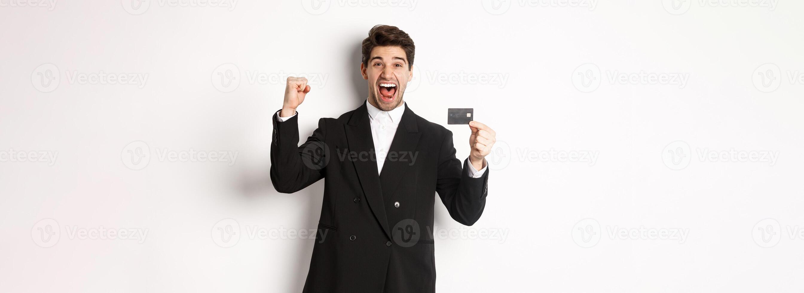 Portrait of excited handsome businessman in suit, rejoicing and showing credit card, standing against white background photo