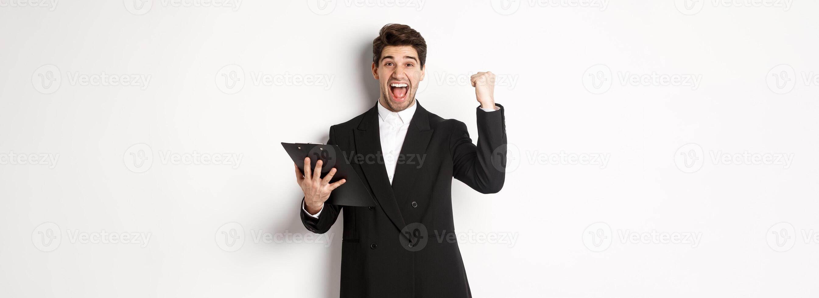 Portrait of excited handsome businessman in black suit, holding clipboard and making fist pump, achieve goal and rejoicing, standing over white background photo
