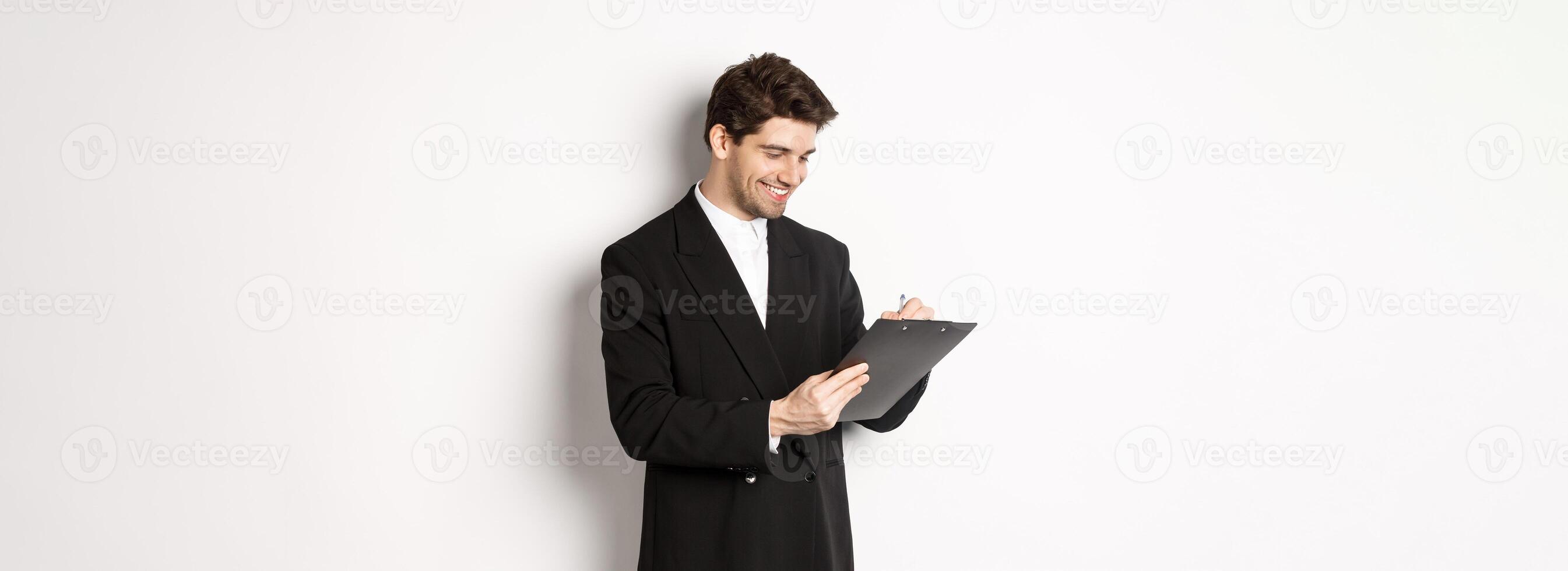 Image of handsome businessman in suit signing documents, looking at clipboard and smiling, standing against white background photo