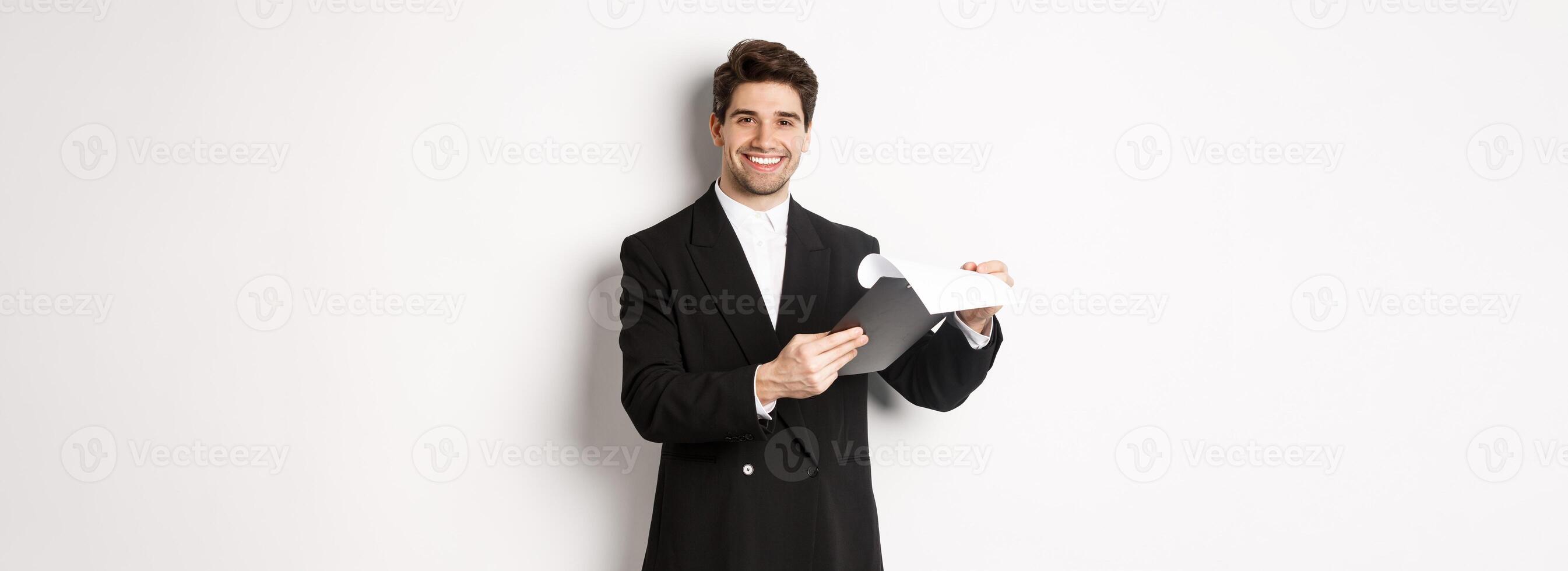 Image of confident businessman in suit looking at documents, holding clipboard and smiling pleased, standing against white background photo