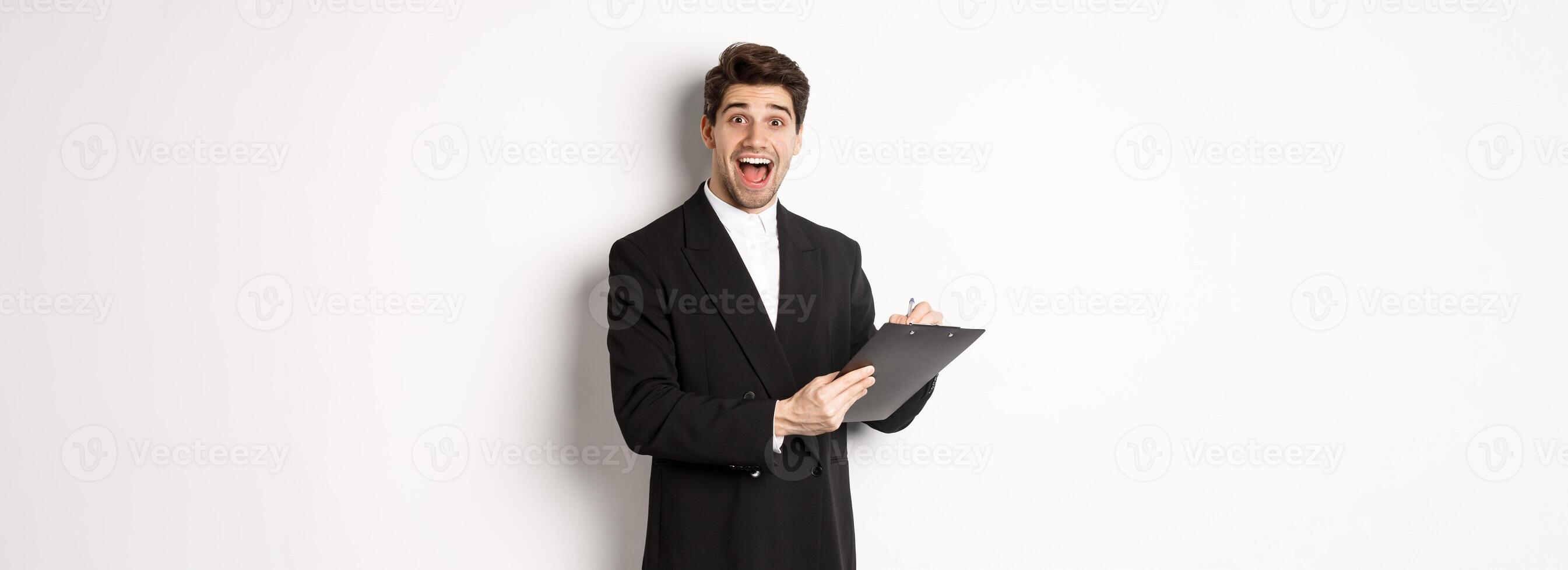 Portrait of handsome businessman in black suit, looking amazed while signing documents on clipboard, standing against white background photo