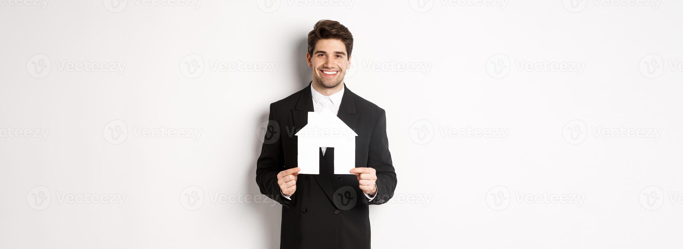 Image of handsome real estate agent in black suit showing home maket, smiling at camera, standing against white background photo