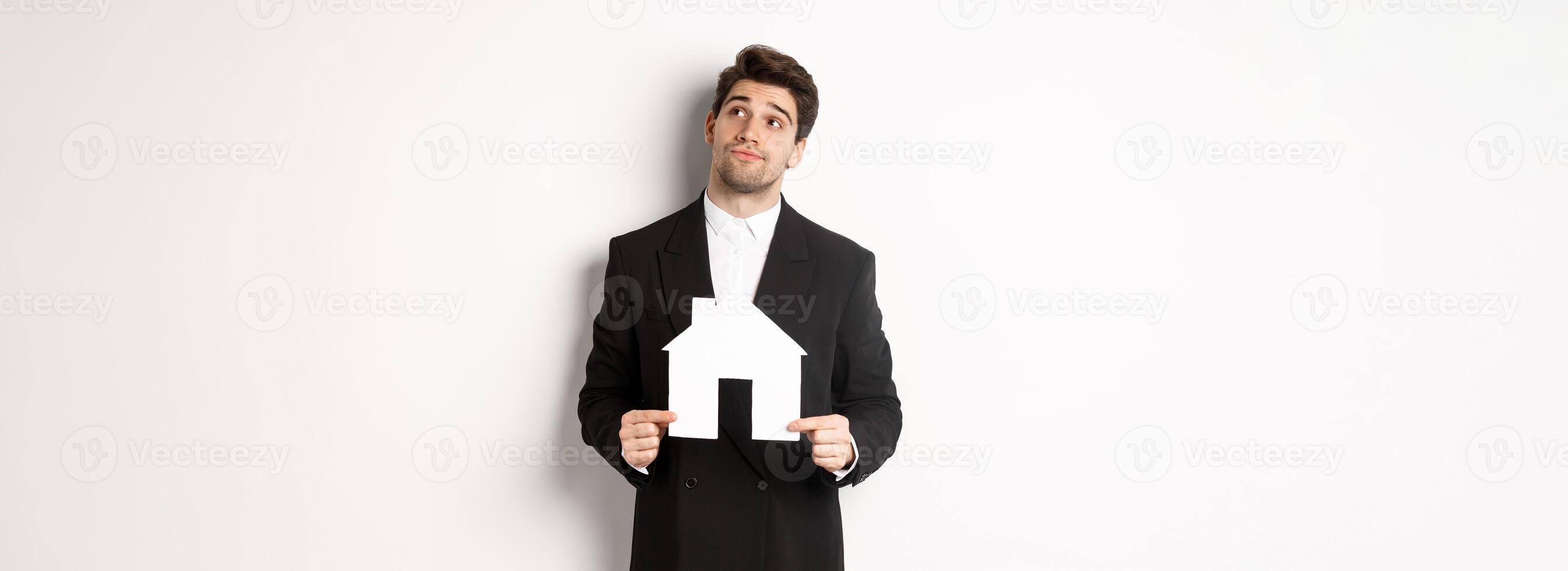 Image of handsome businessman in black suit, looking for home, holding house maket and gazing dreamy at upper right corner, standing against white background photo