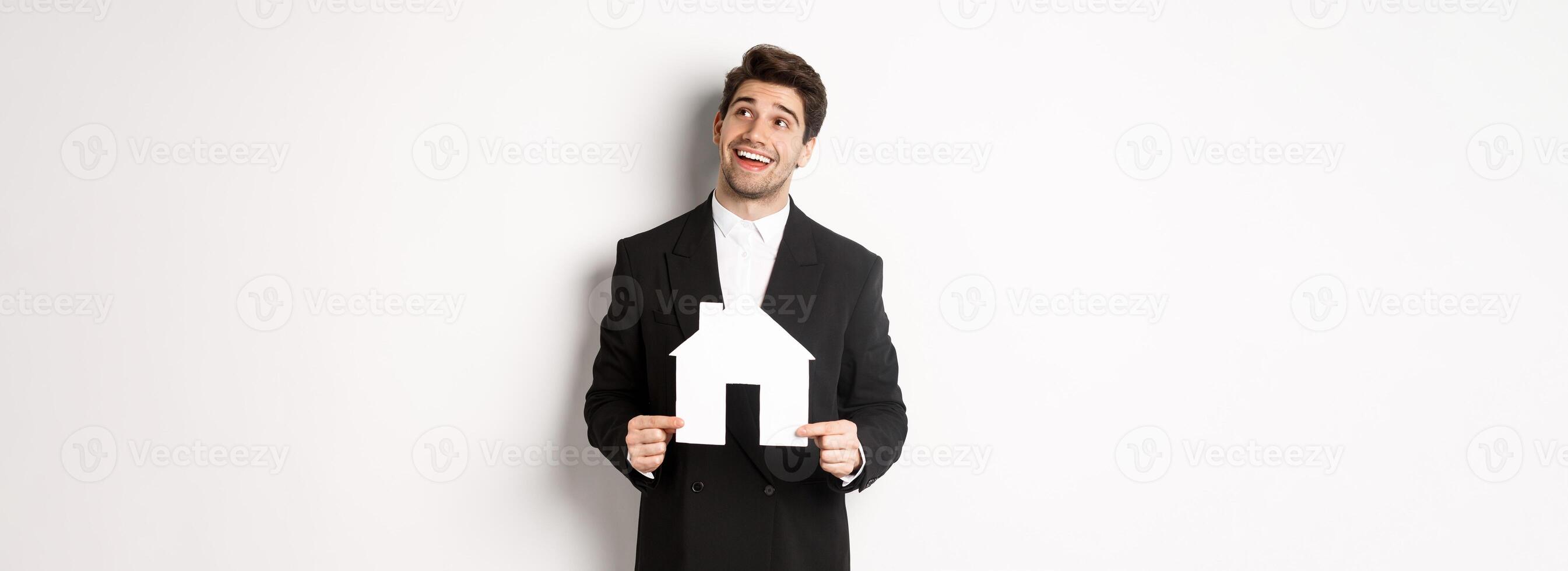 Portrait of handsome man in suit searching for home, holding paper house and looking at upper right corner dreamy, standing over white background photo