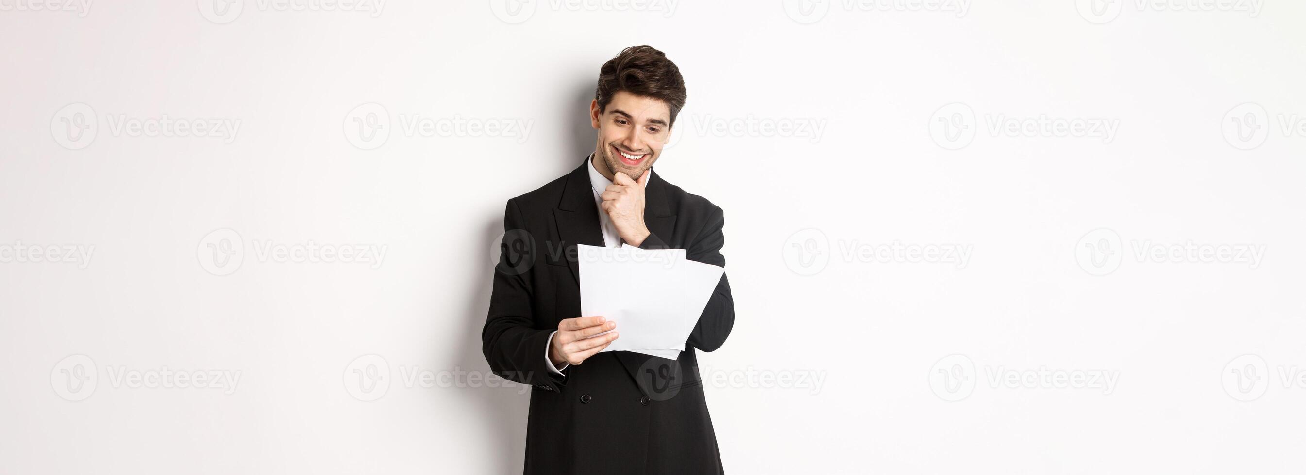 Image of handsome businessman in black suit, looking pleased at documents, reading report and smiling, standing against white background photo