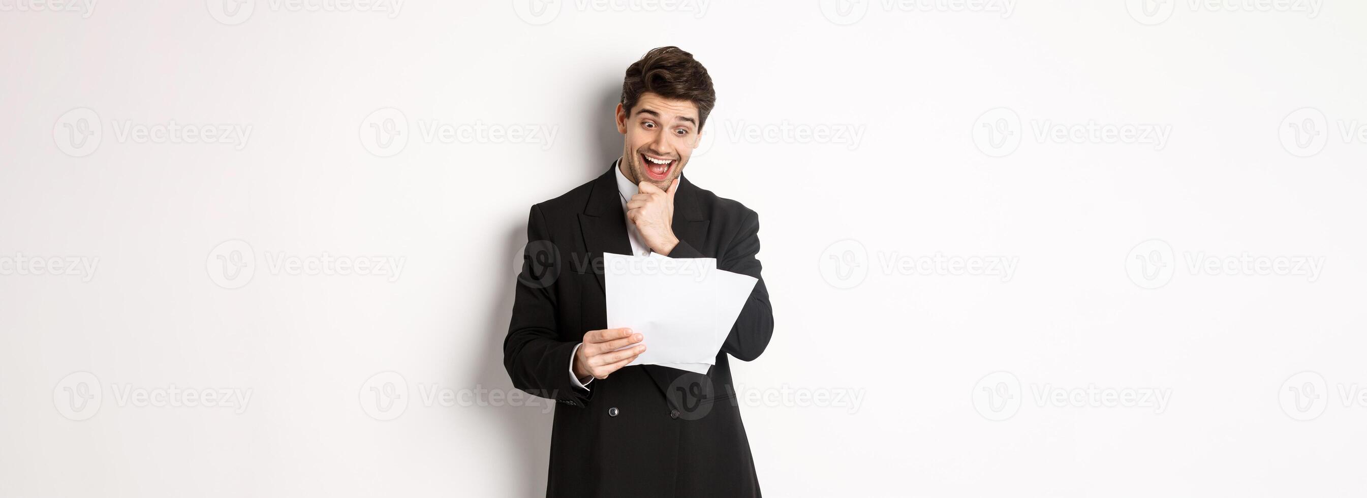 Portrait of handsome businessman looking excited at documents, working, standing against white background in black suit photo