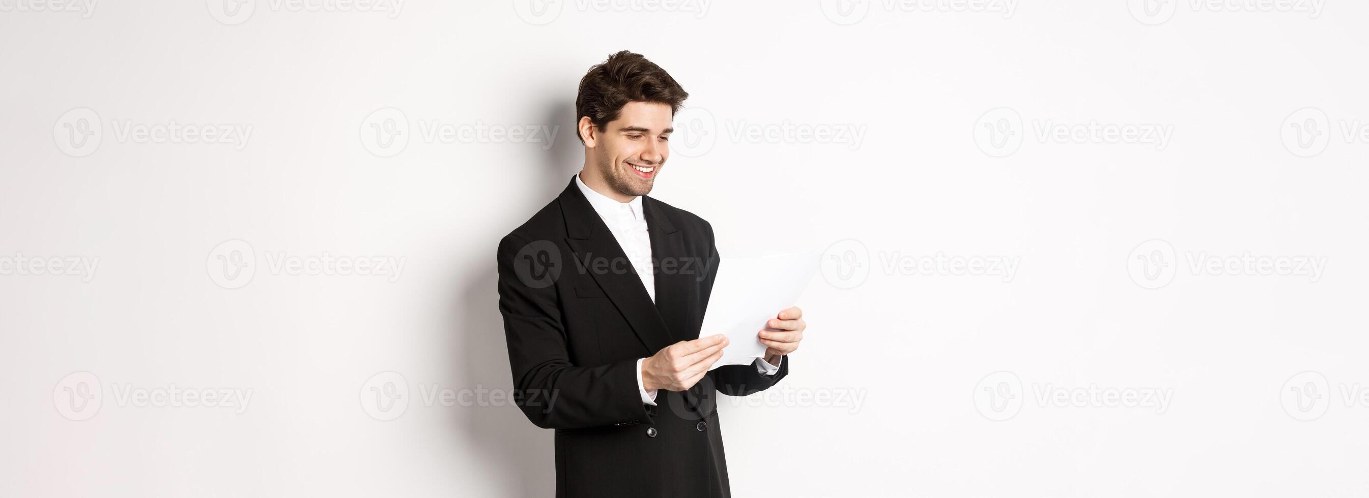Image of attractive businessman in black suit, reading document and smiling, working on report, standing against white background photo
