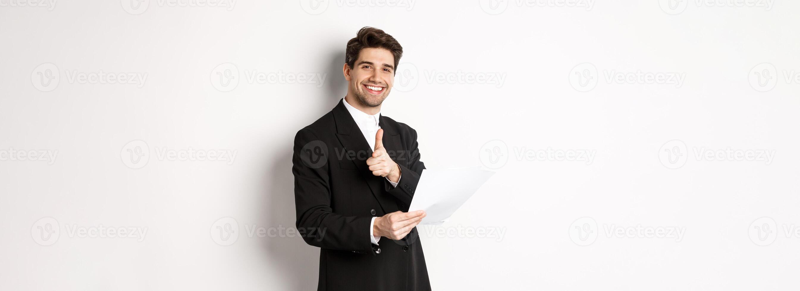 Image of handsome businessman in black suit, holding document and pointing finger at camera, praising good job, standing against white background photo
