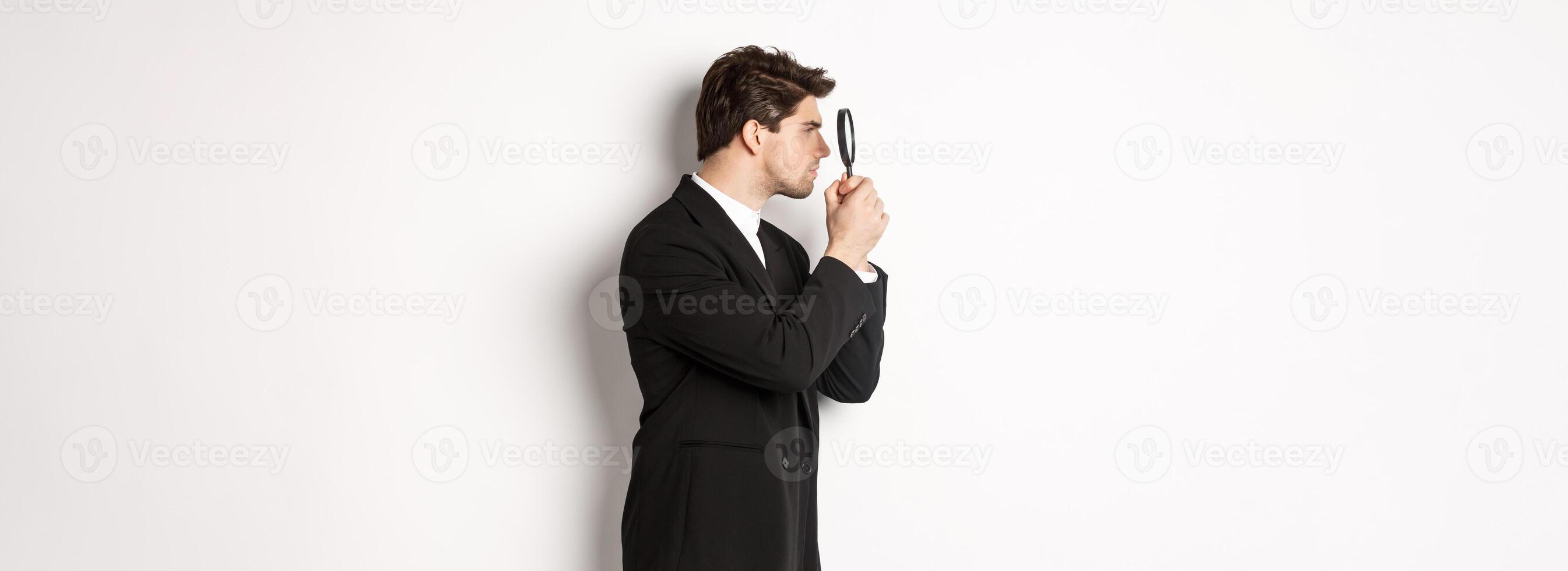 Profile shot of handsome businessman in black suit, looking through magnifying glass and searching for something, standing over white background photo