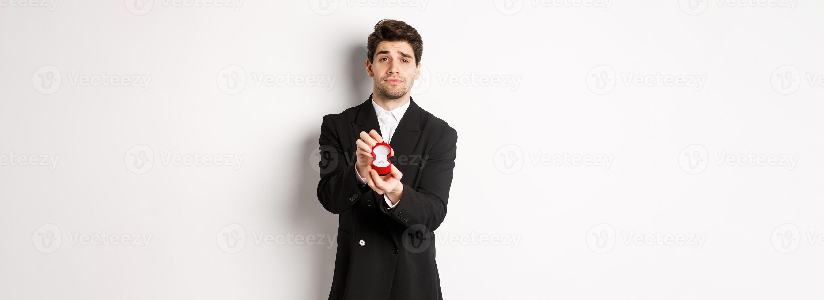 Portrait of handsome man making a proposal, asking to marry him, showing ring, standing in black suit against white background photo