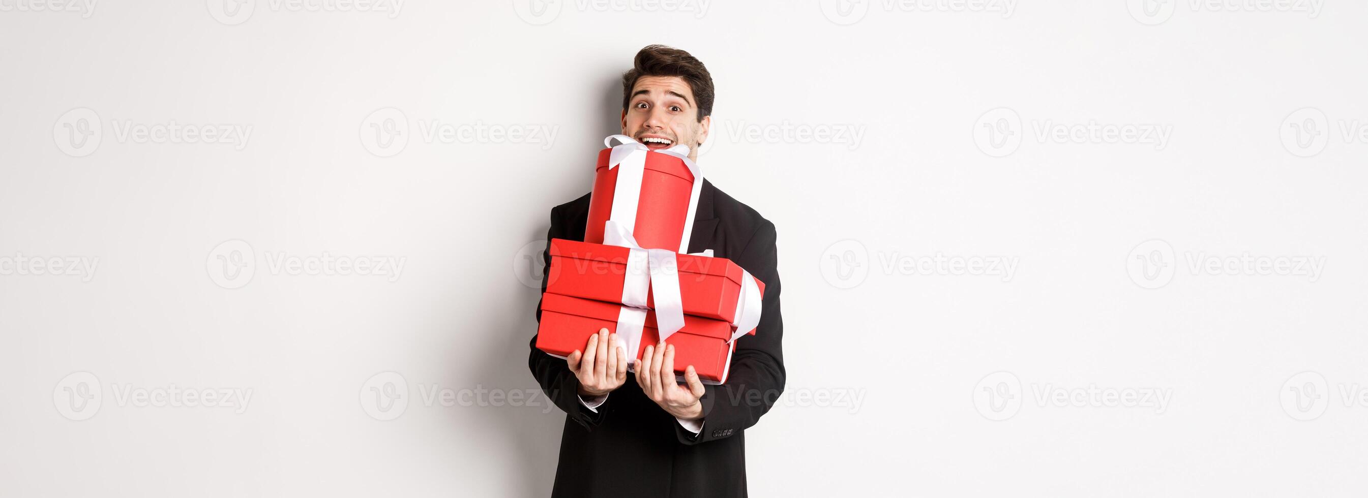 Concept of christmas holidays, celebration and lifestyle. Image of happy man in suit carry presents for new year, holding boxes with gifts and smiling, standing against white background photo