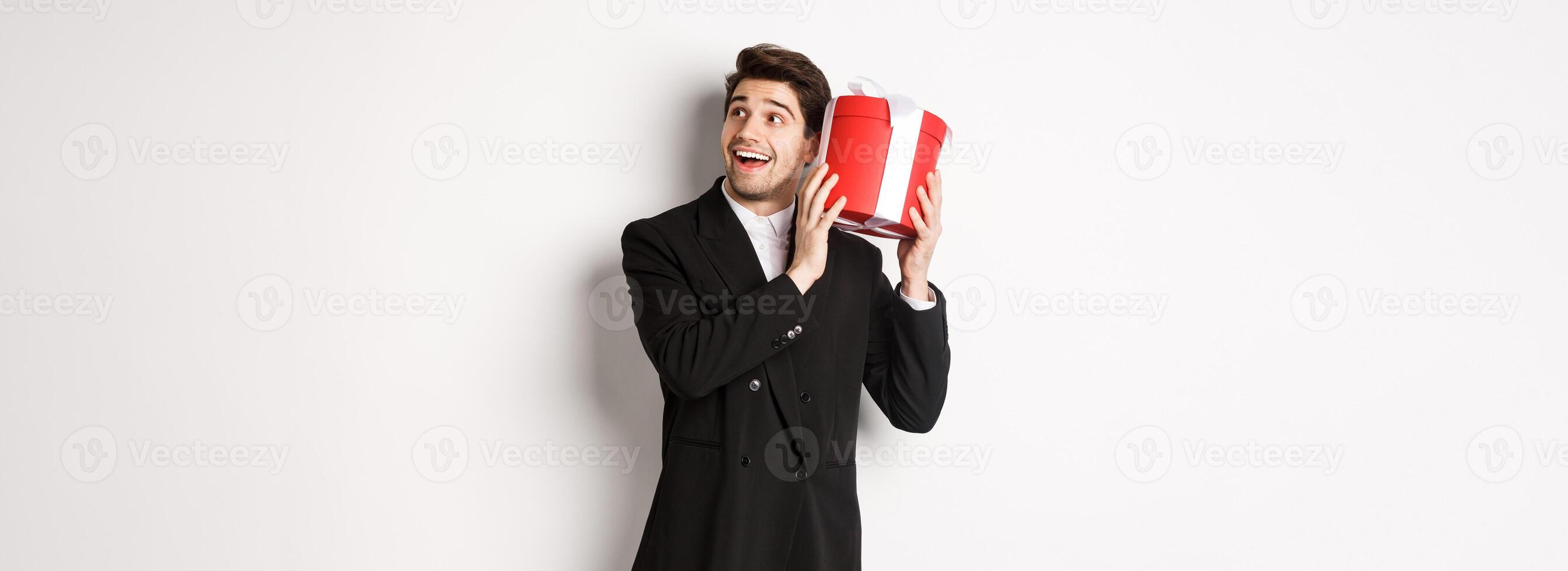Concept of christmas holidays, celebration and lifestyle. Image of attractive man in black suit, shaking present to guess what inside, enjoying new year, standing against white background photo