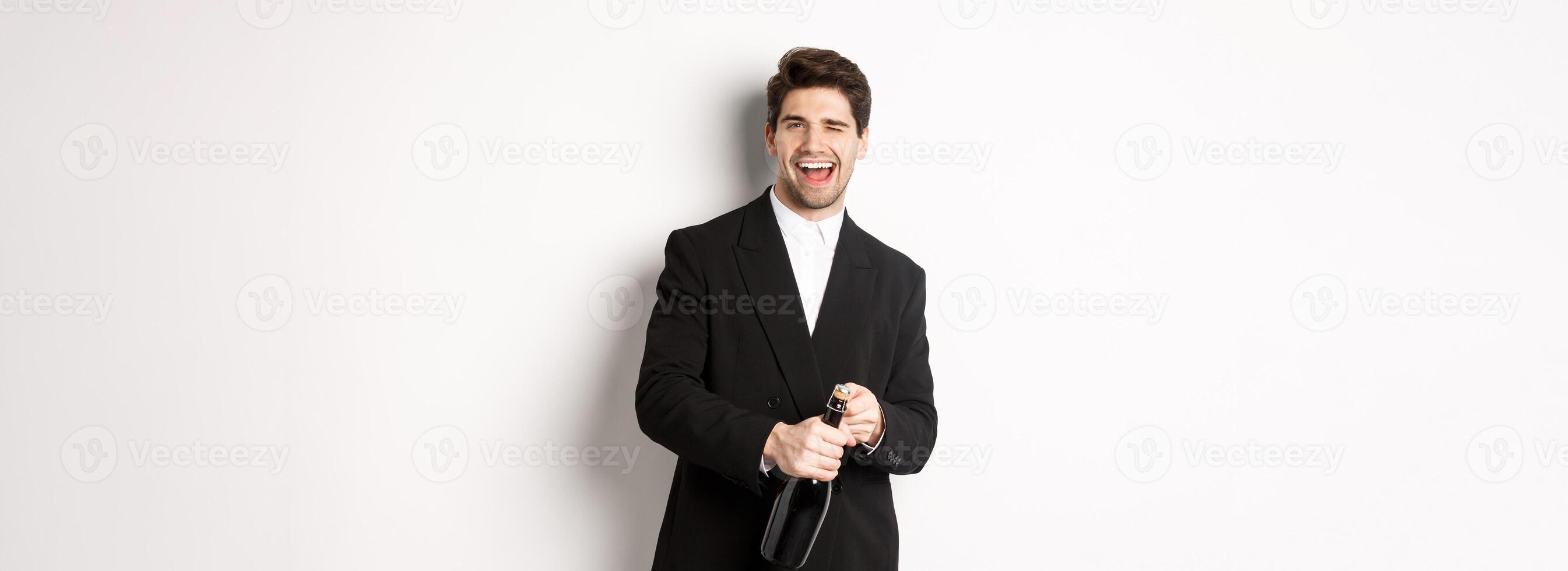Image of handsome, confident man in black suit, celebrating holiday, open a bottle of champagne and partying, standing joyful against white background photo