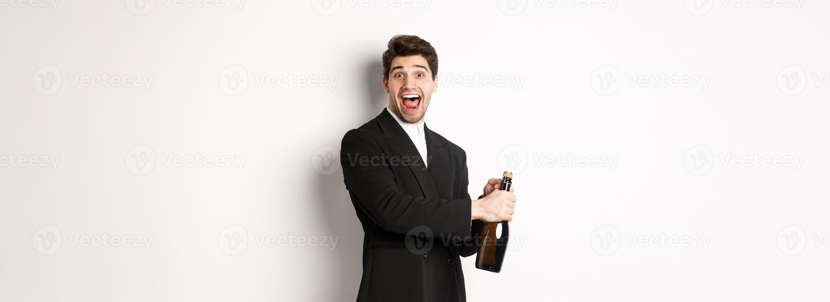 Portrait of attractive man in black suit, winking at camera and opening bottle of champagne, celebrating new year, standing against white background photo