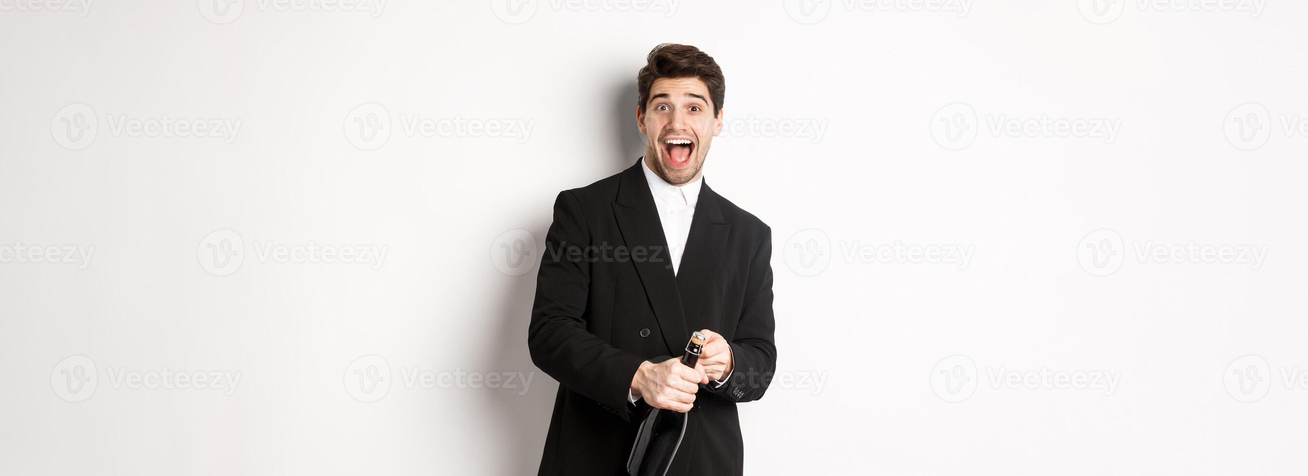 Image of attractive man in black suit having a party, celebrating new year and opening bottle of champagne, standing happy against white background photo