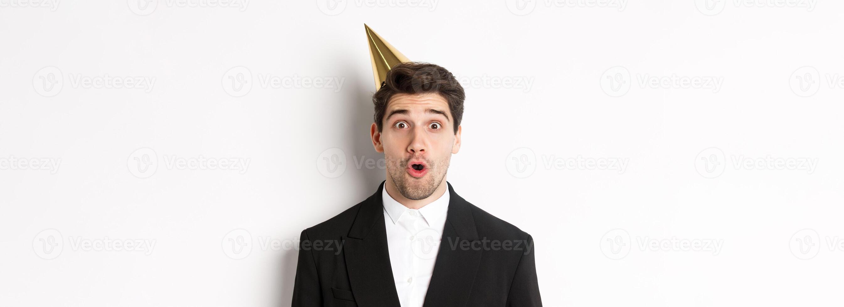 Close-up of attractive man in party hat and trendy suit, looking surprised, celebrating new year, standing against white background photo