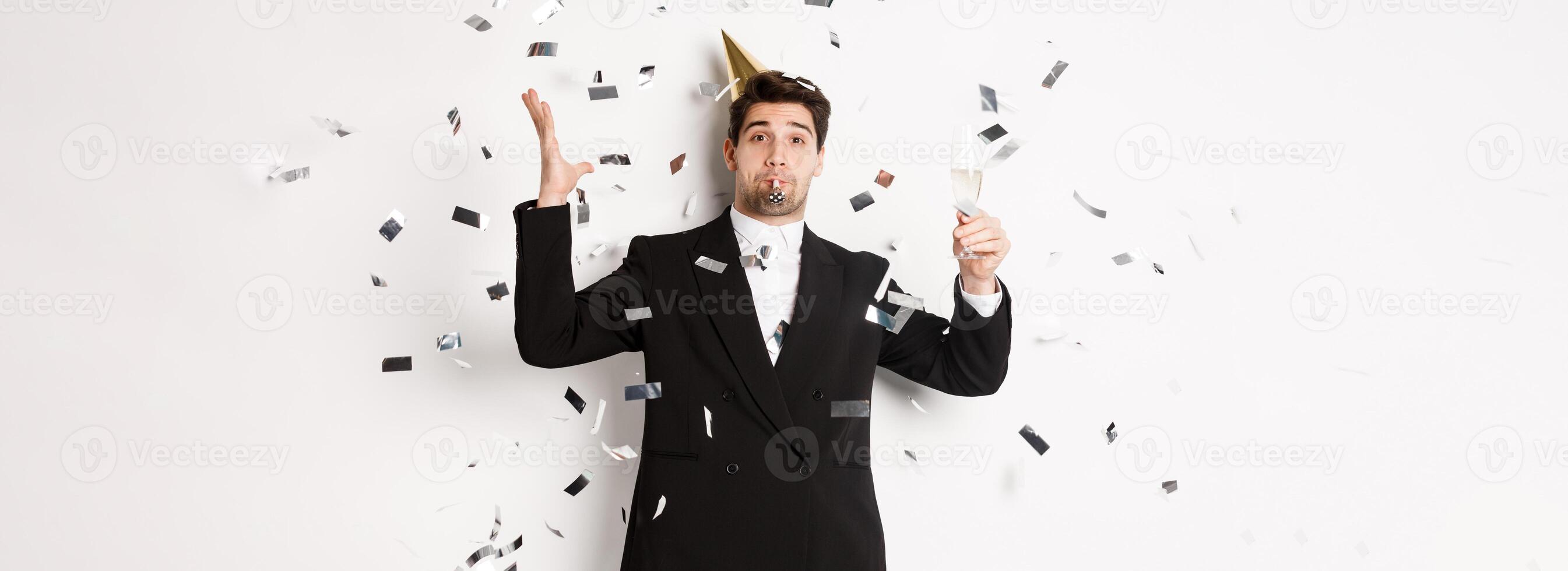 Handsome party guy in black suit having fun, celebrating new year, blowing whistle and drinking champagne while confetti falling, wishing happy holidays, standing against white background photo