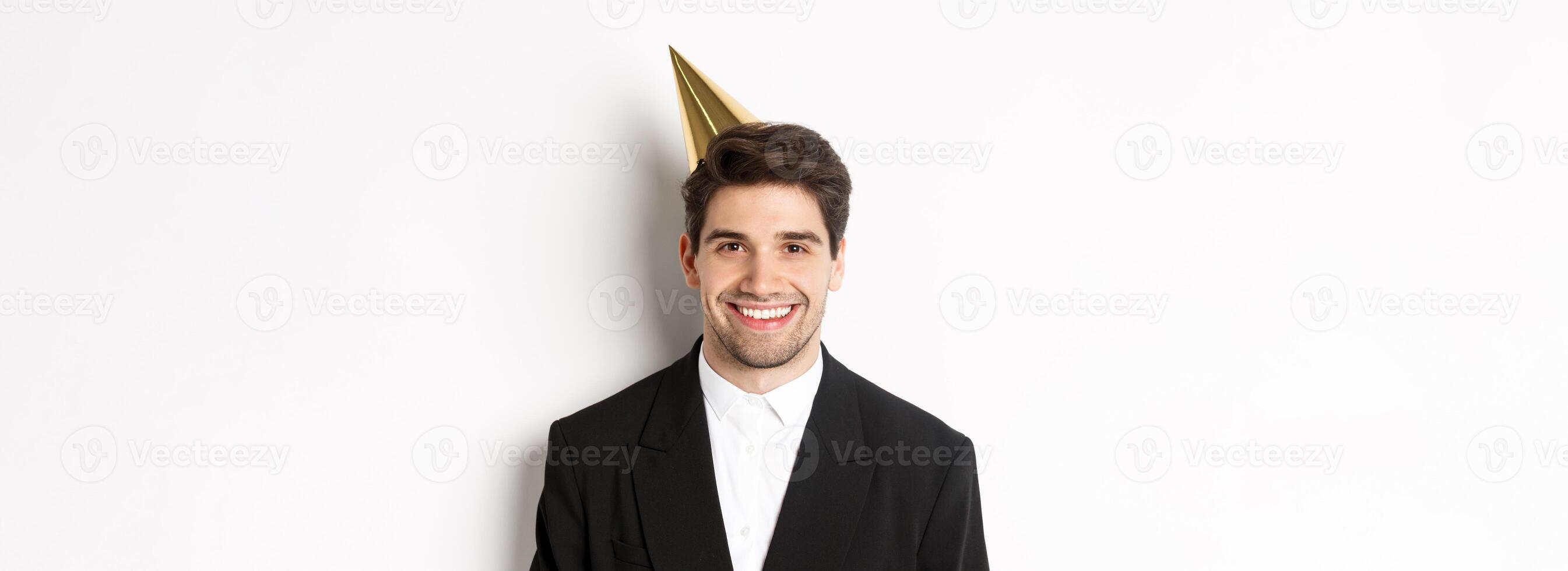 Close-up of handsome happy man in suit and party hat, smiling joyful, celebrating holiday, standing against white background photo
