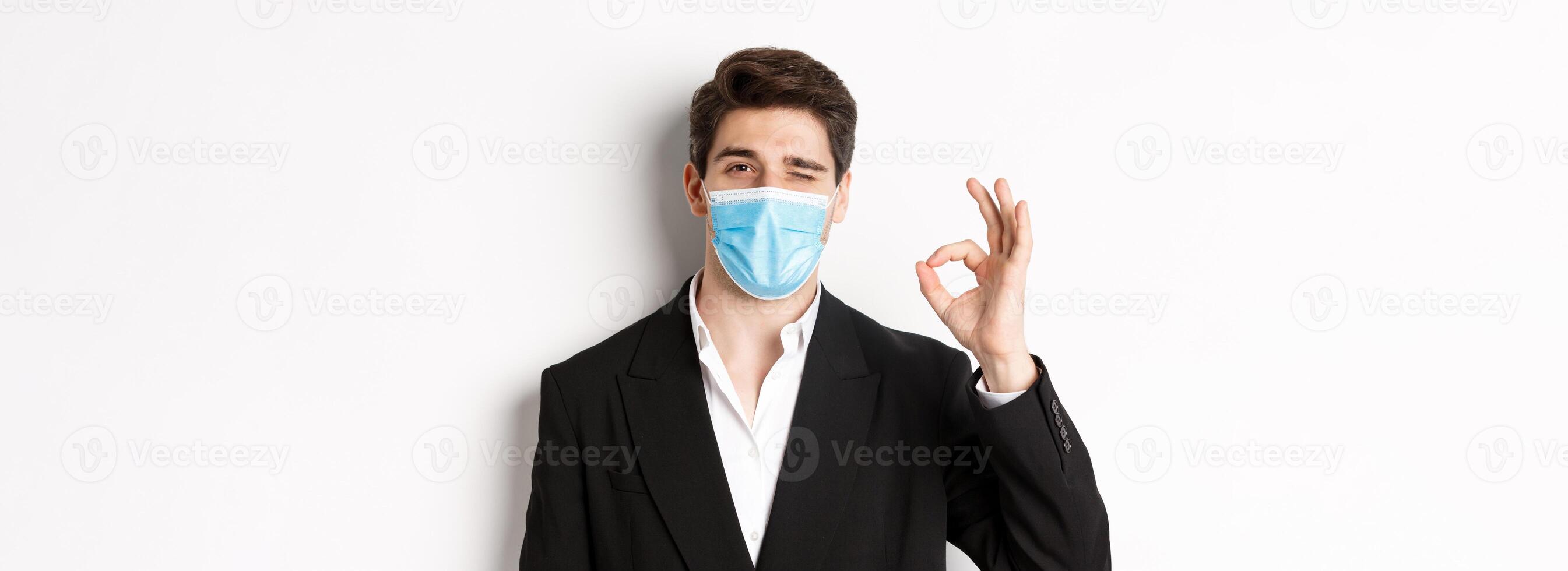 Concept of covid-19, business and social distancing. Close-up of good-looking businessman in trendy suit and medical mask, showing okay sign and winking, standing against white background photo