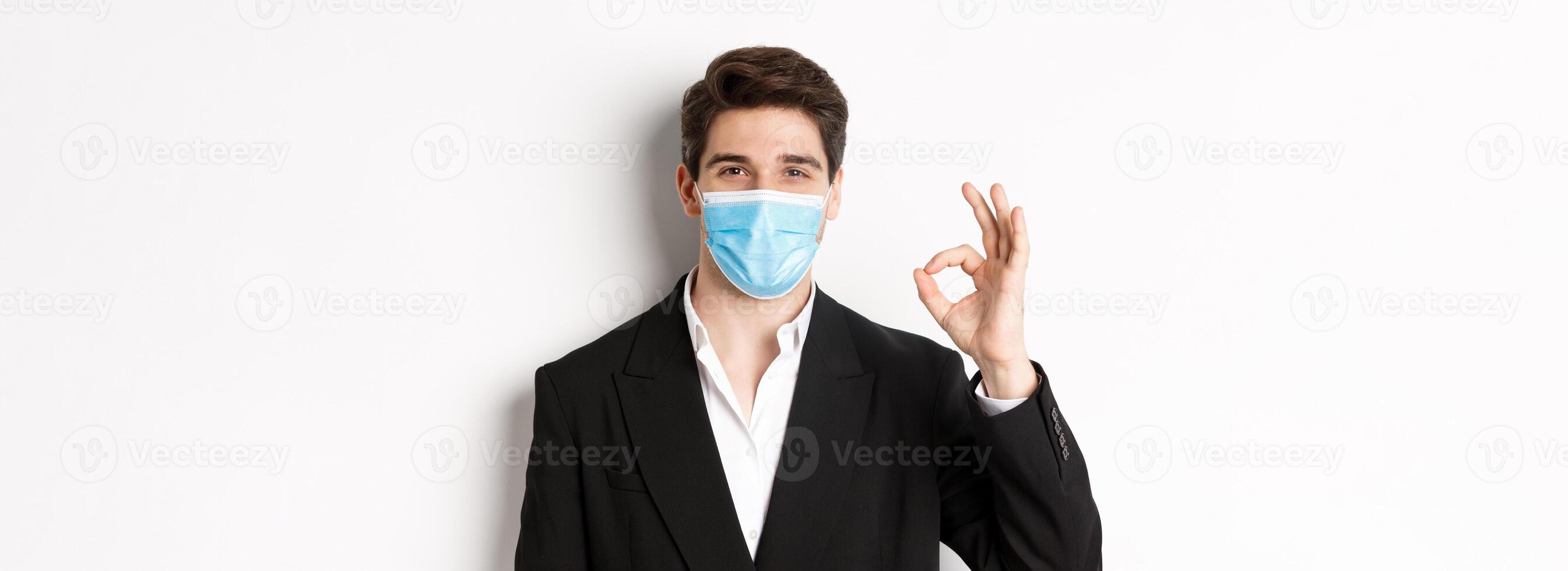 Concept of covid-19, business and social distancing. Close-up of good-looking businessman in trendy suit and medical mask, showing okay sign, recommending something, white background photo